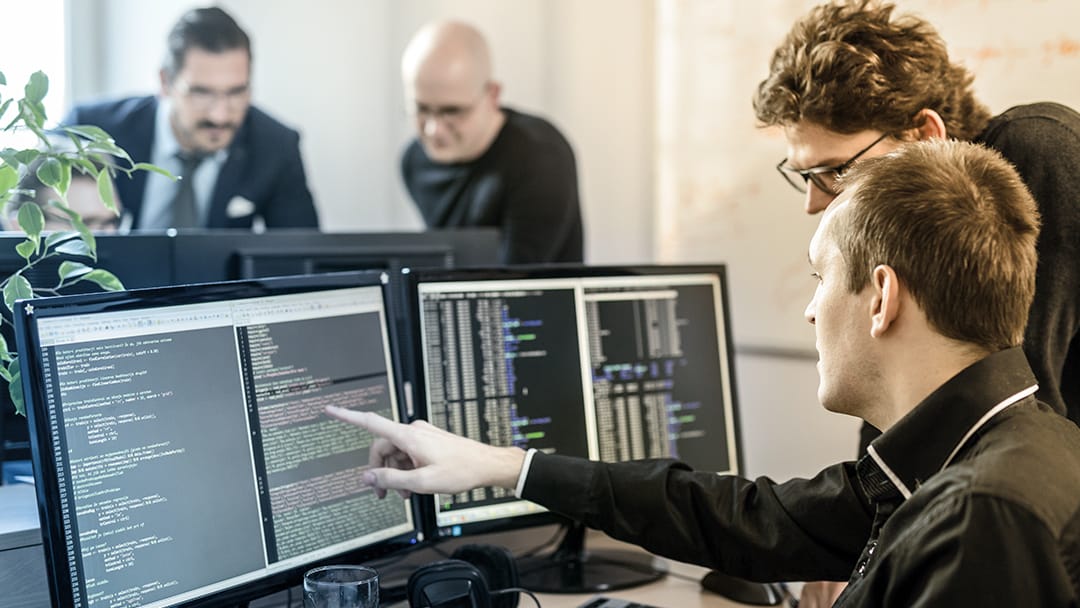 一名技术人员指着同事桌面电脑屏幕上的软件代码。